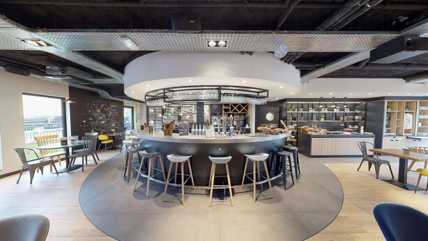 A modern bar area with a circular bar, stools, and a kitchen in the background. - Campanile Fontainebleau (Fontainebleau)