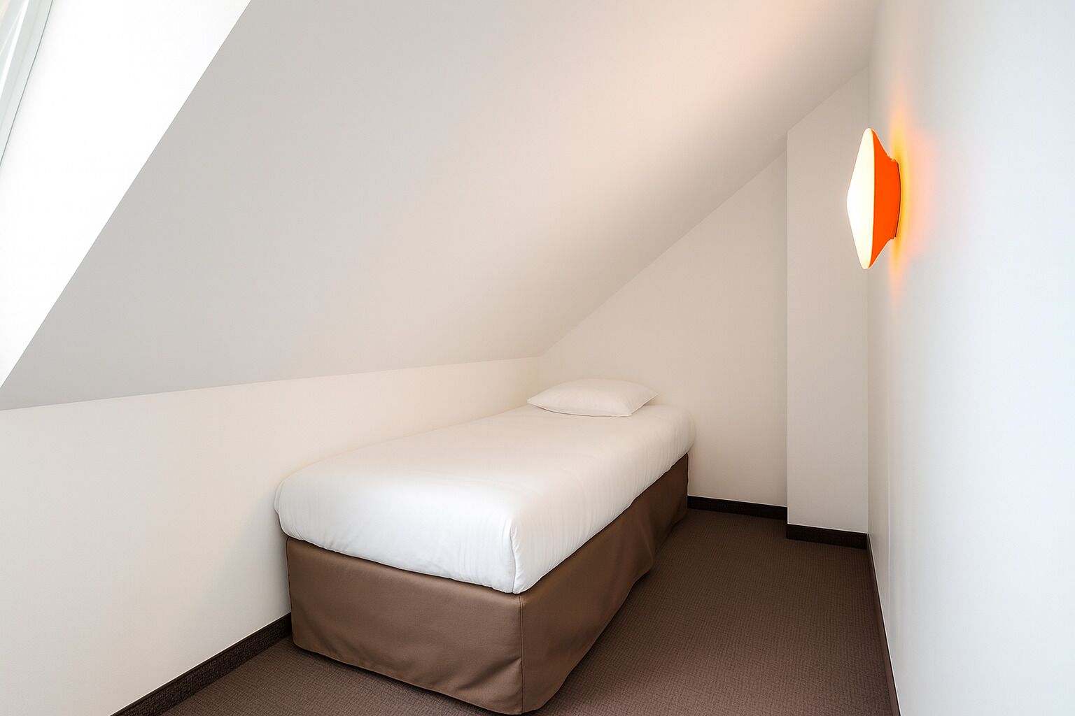 A small, single-bed room with a sloped ceiling, a wall-mounted light, and a carpeted floor.