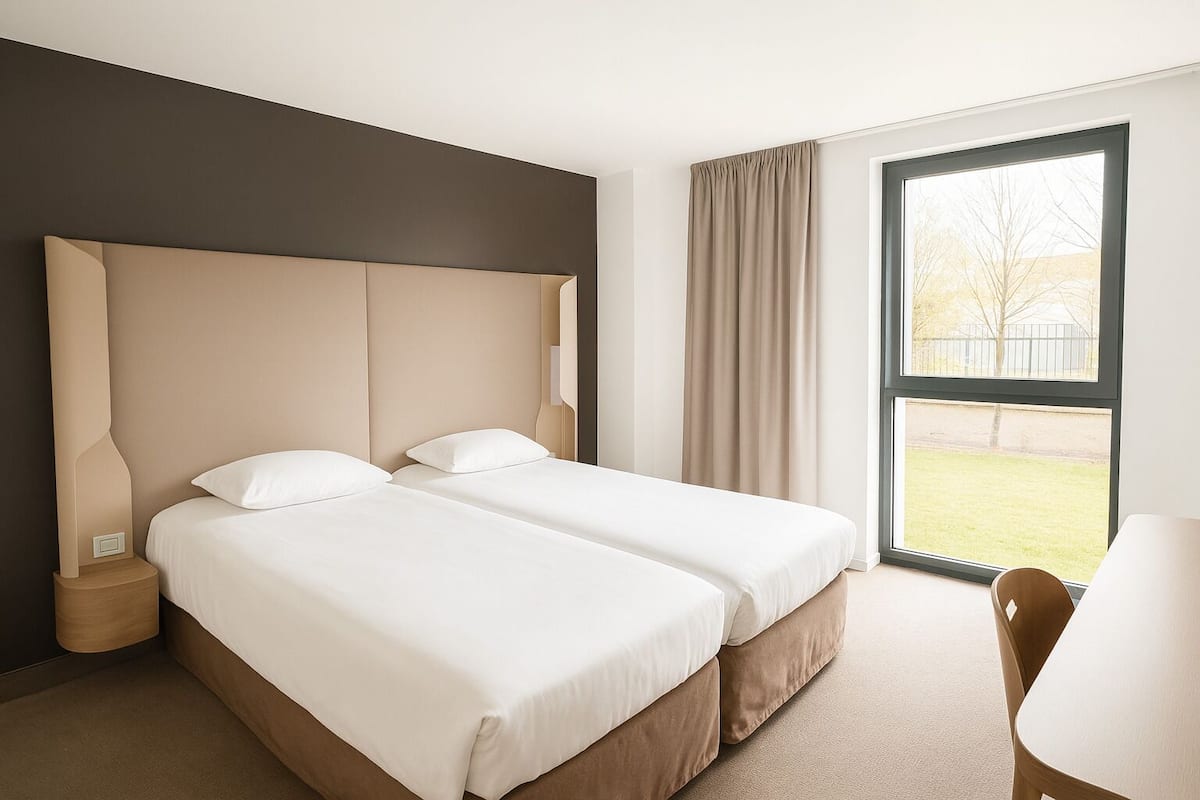 A hotel room with two beds, a large window, a chair, and a lamp.