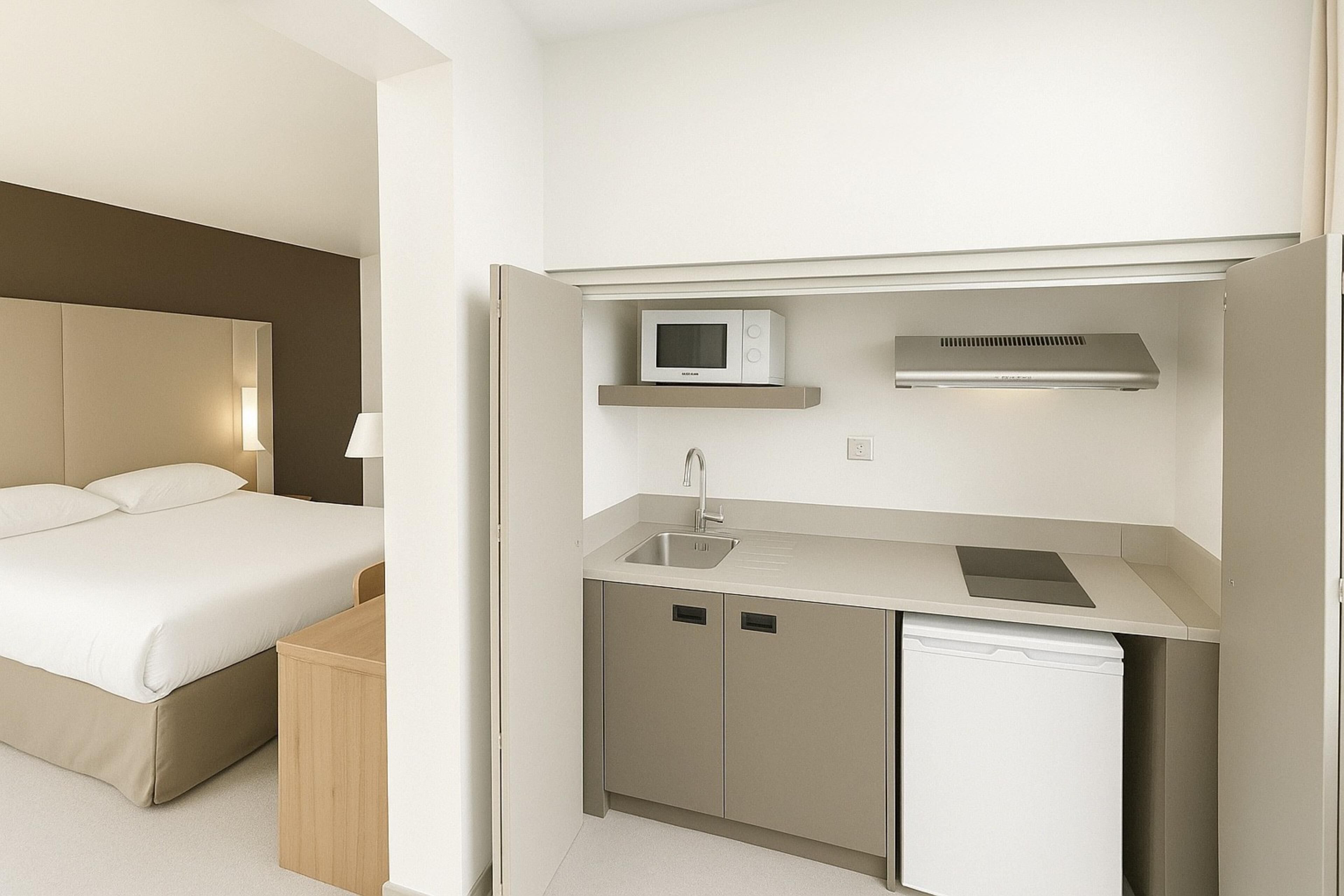 Habitación de hotel compacta con cama, pequeña cocina y mesita de noche.