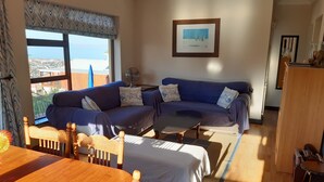 A cozy living room with a blue sofa, a dining table, and a view of the sea. - A Place in Thyme (Cape Town)