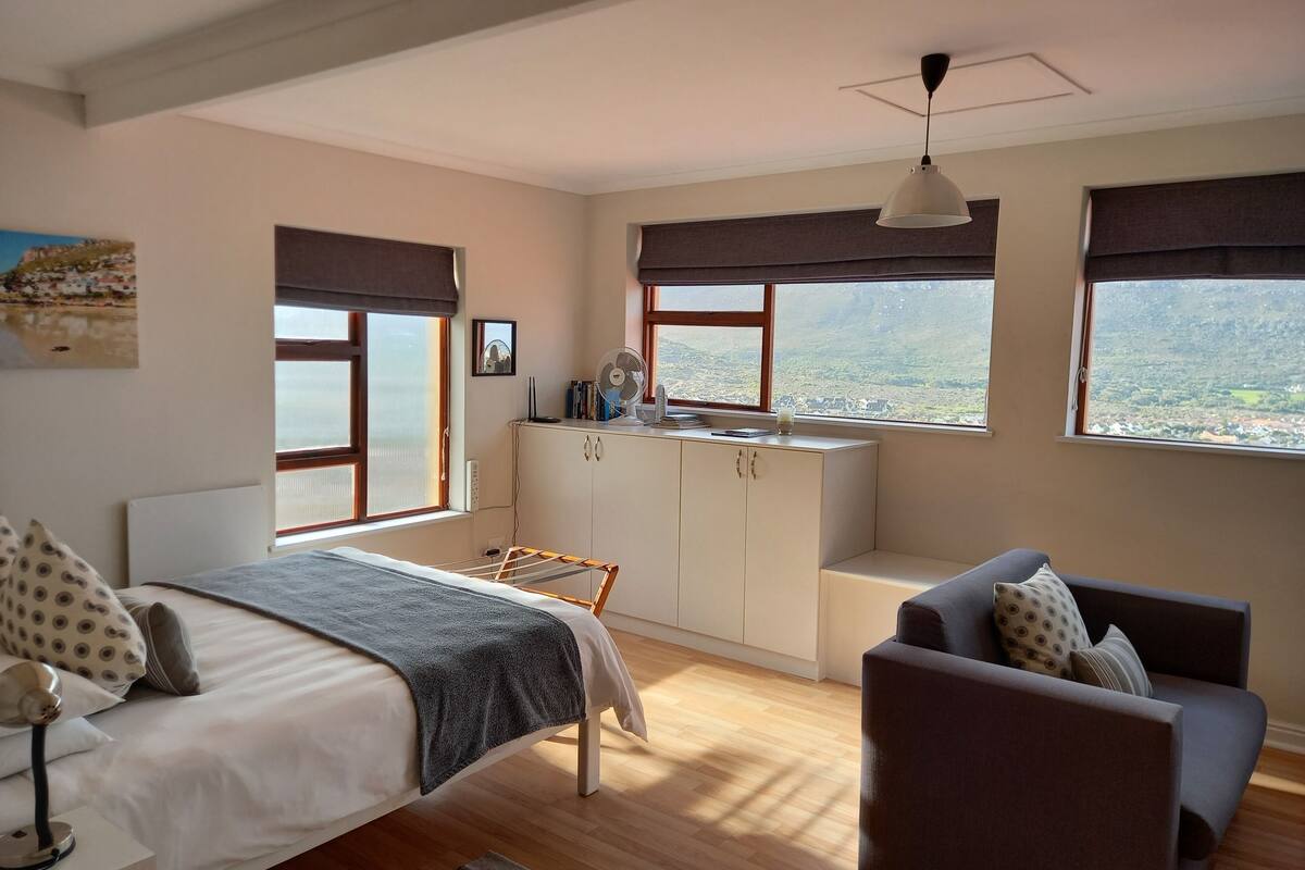 A bedroom with a bed, a desk, a chair, and a view of the landscape through large windows.