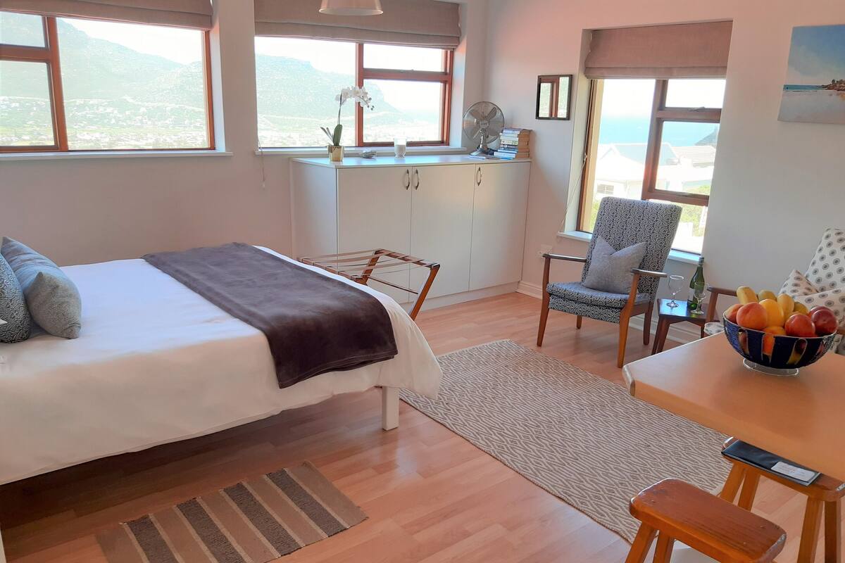 A bedroom with a bed, a desk with a fruit bowl, a chair, and a view of mountains through large windows.