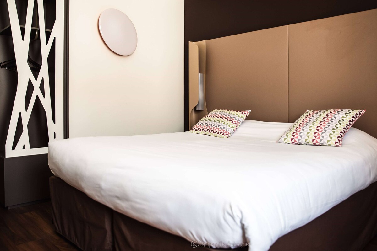 Een moderne hotelkamer met een groot bed, twee gestreepte kussens, een nachtkastje met een lamp en een decoratief wandpaneel.