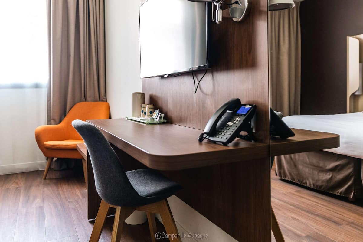 A hotel room with a wooden desk, a chair, a bed, a TV, a phone, and a window with curtains.