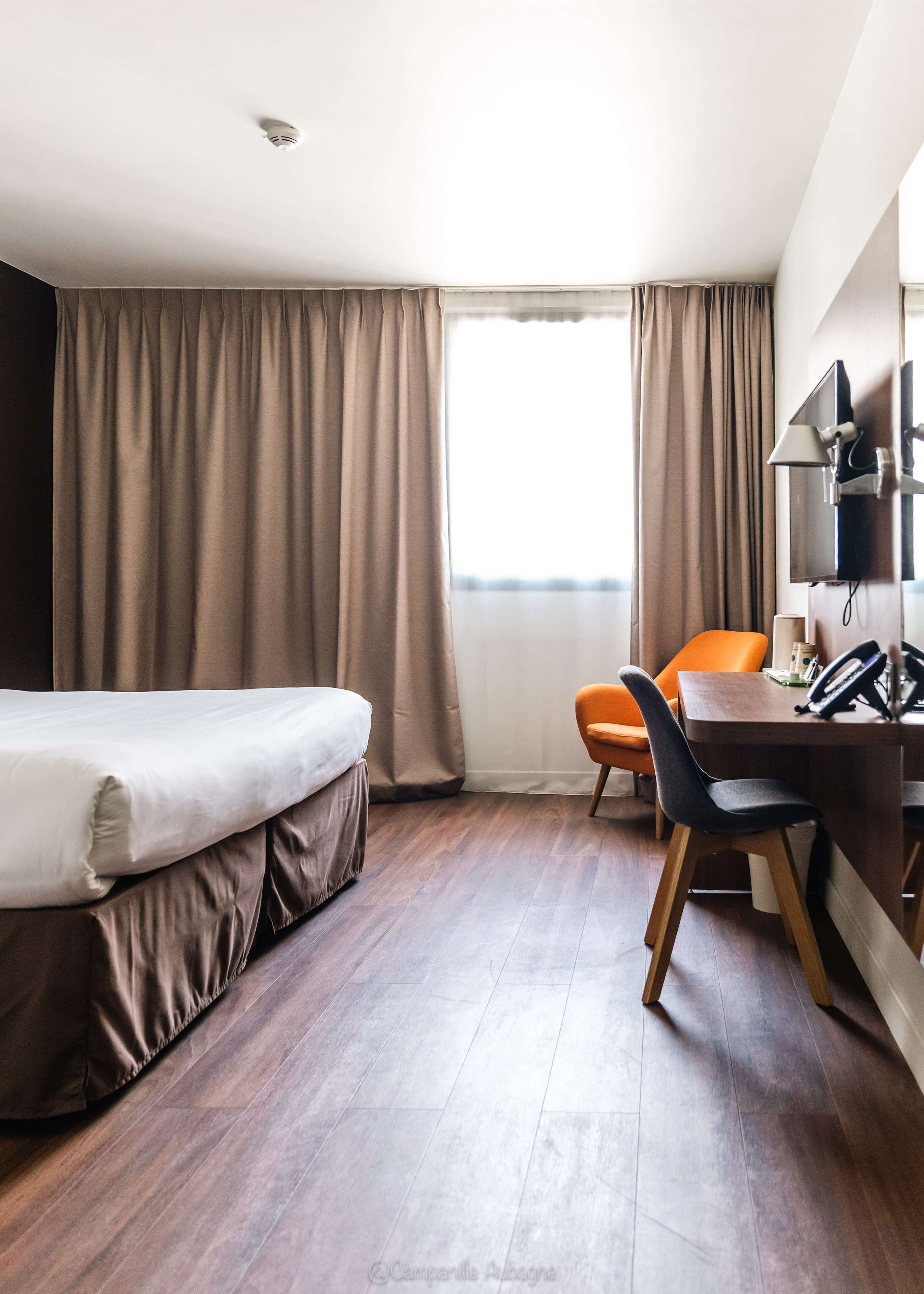 Une chambre d’hôtel avec un lit, un bureau, une chaise orange, une télévision et une fenêtre avec des rideaux.