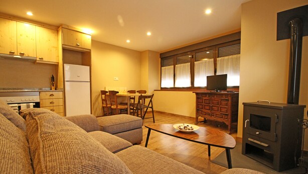 A living room with a sofa, a coffee table, and a television. There is a kitchen area with wooden cabinets and a refrigerator. - Apartamentos Mont Romies Plaza (Naut Aran)