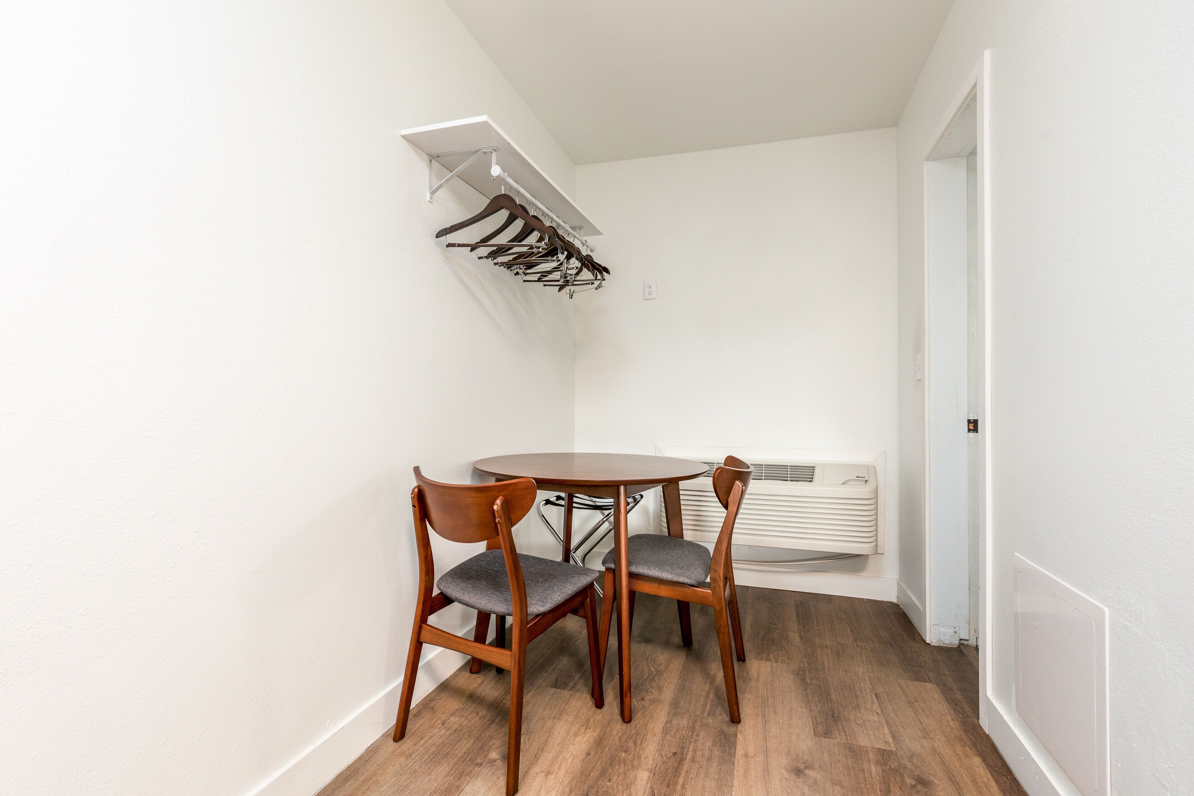 Un pequeño comedor con una mesa redonda y cuatro sillas de madera, una pared blanca y un equipo de aire acondicionado empotrado.