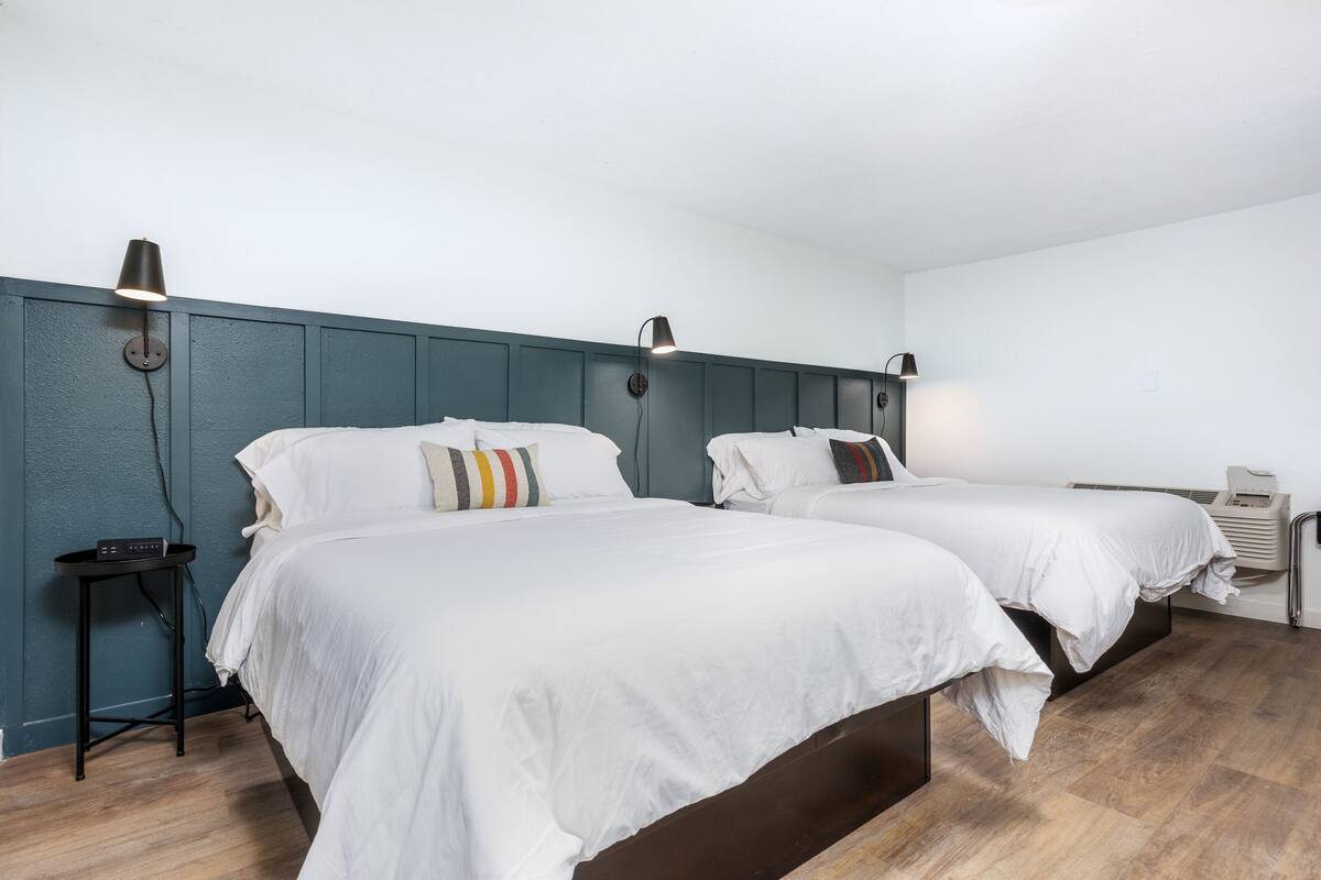 A hotel room with two beds, each with white bedding and a colorful pillow. There are wall-mounted lamps and a small table with a radio.