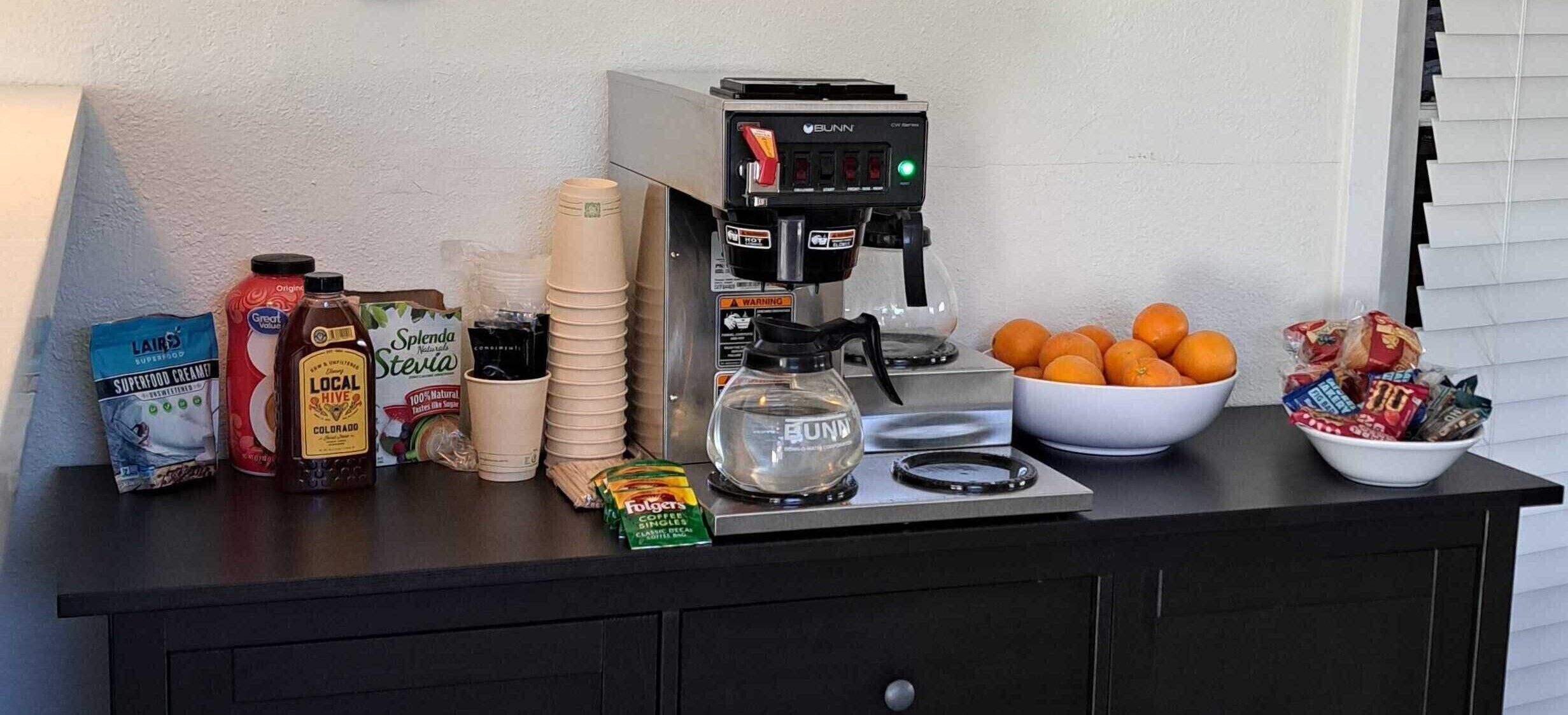 A kitchen counter with a coffee maker, stacks of paper cups, a bowl of oranges, and various food items.