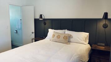 A neatly made bed with white linens and a decorative pillow, a bedside table with a lamp, and a door with a towel hanging on it.
