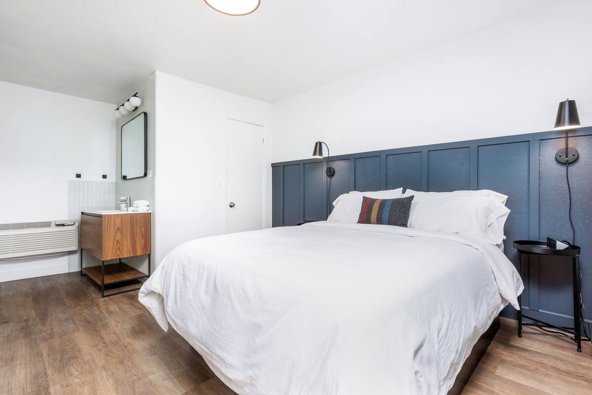 A bedroom with a large bed, a bedside table, a nightstand, a mirror, and a wall-mounted lamp.