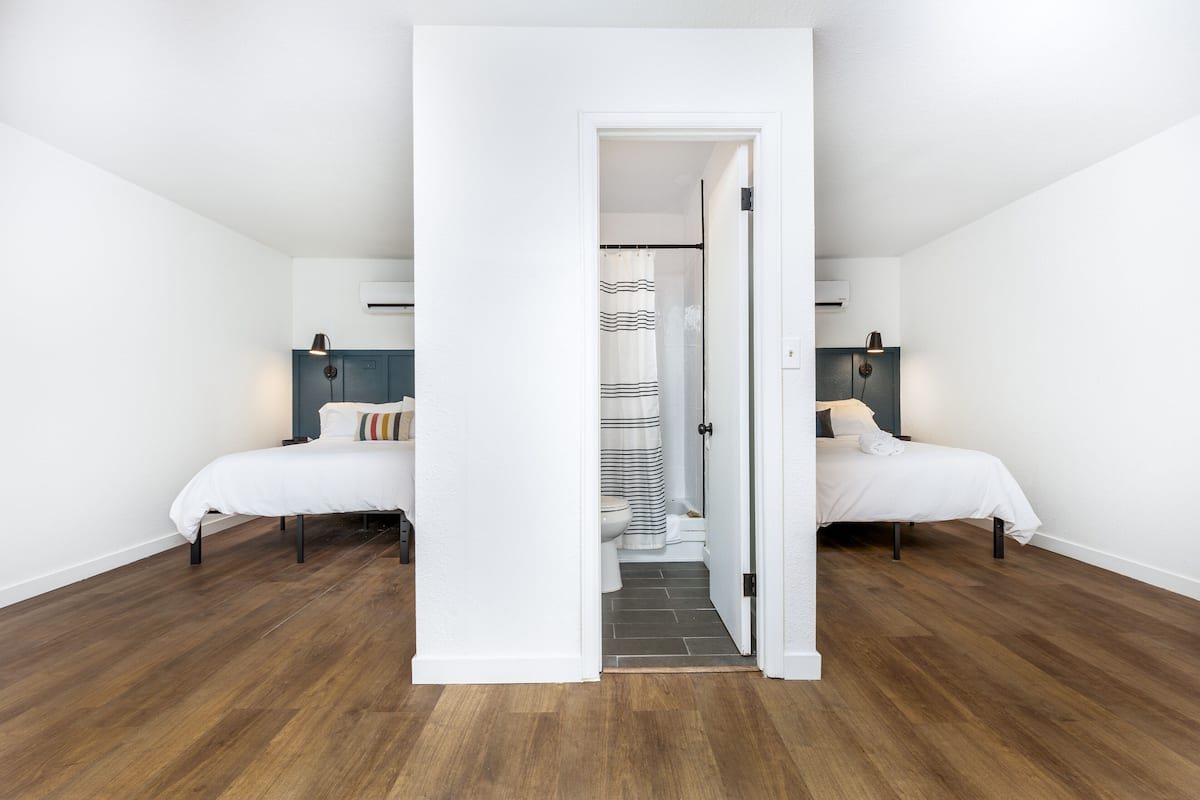 A bedroom with two beds, a bathroom with a shower curtain, and a hallway leading to another room.
