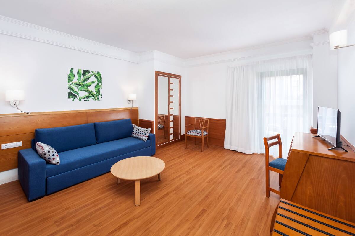 Una camera d'albergo moderna con un divano blu, pavimento in legno, un piccolo tavolino rotondo, una scrivania in legno con una sedia e un grande specchio.