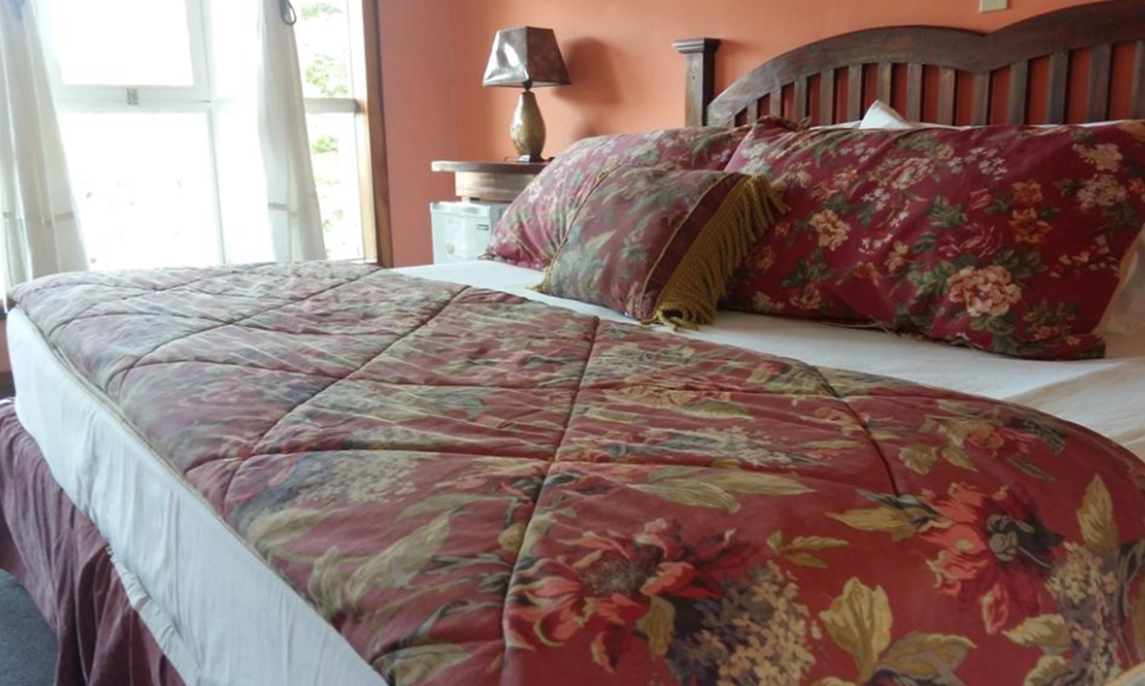 A bed with a floral bedspread and pillows, a wooden headboard, a bedside lamp, and a window with curtains.