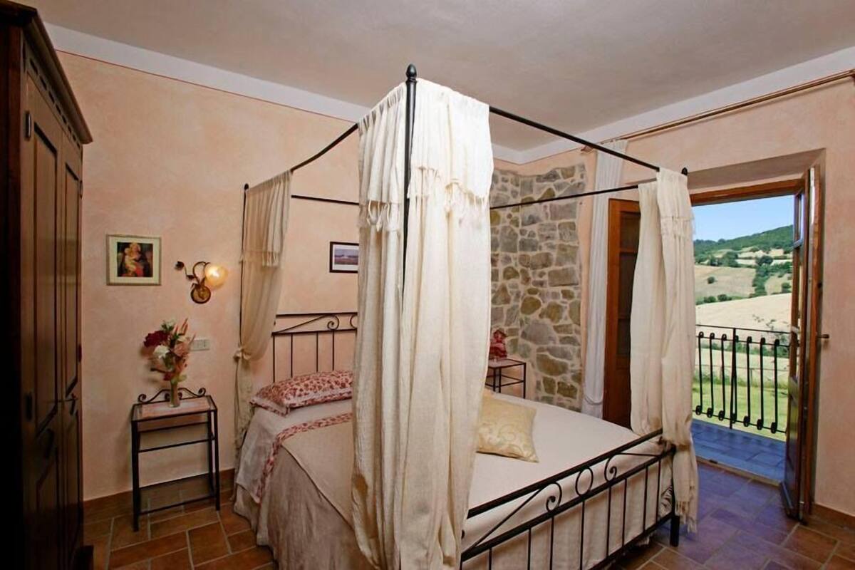 Une chambre à coucher avec un lit à baldaquin, une petite table de chevet ornée d’un vase de fleurs et une vue sur la campagne, visible à travers une porte ouverte.