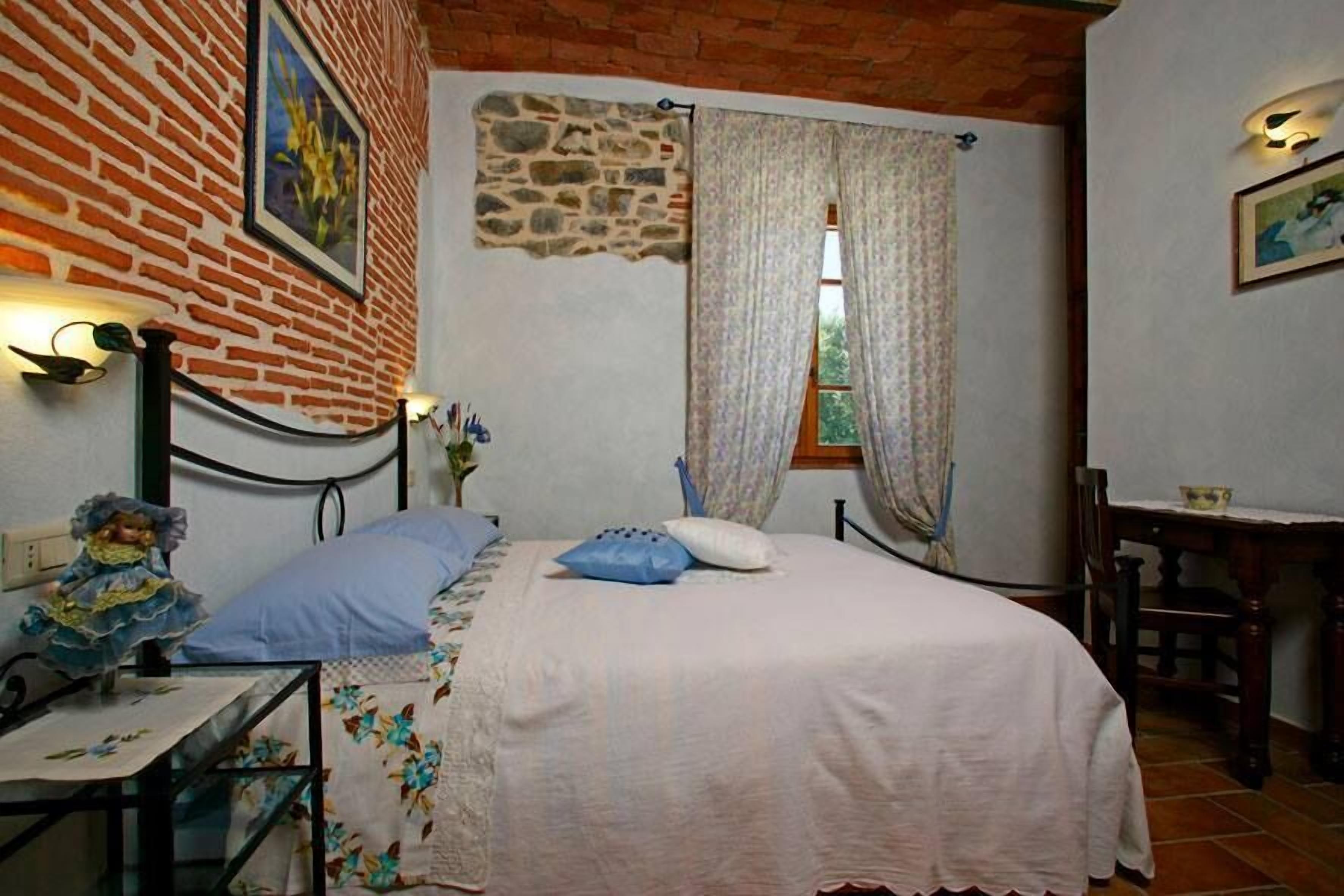 A bedroom with a brick wall, a bed with a white bedspread, a nightstand with a lamp, and a window with curtains.