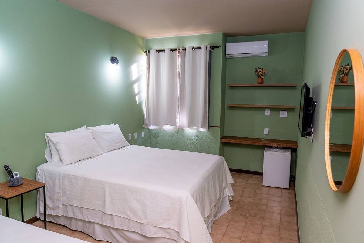 A bedroom with a bed, a bedside table with a phone, a small desk, a mirror, a shelf with decorative items, and a wall-mounted air conditioner.