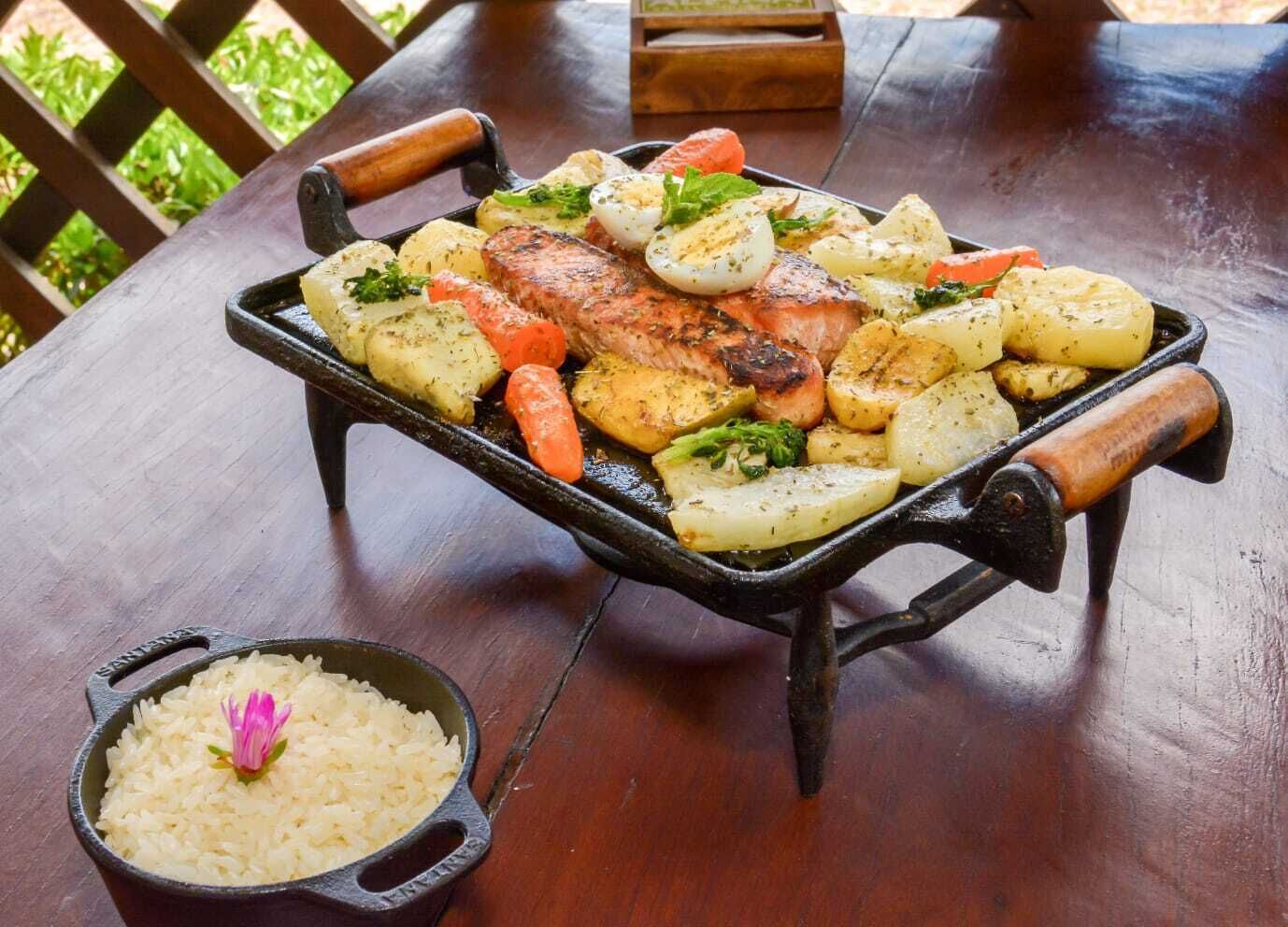 A meal of grilled fish, potatoes, and carrots on a cast iron skillet, with a side of rice in a separate pot.