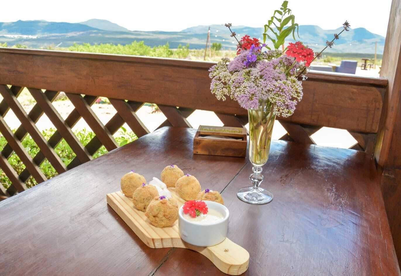 Une table en bois sur laquelle repose un plateau en bois contenant des pâtisseries rondes, un petit bol blanc avec une fleur rouge et un grand vase en verre contenant des fleurs violettes et rouges.