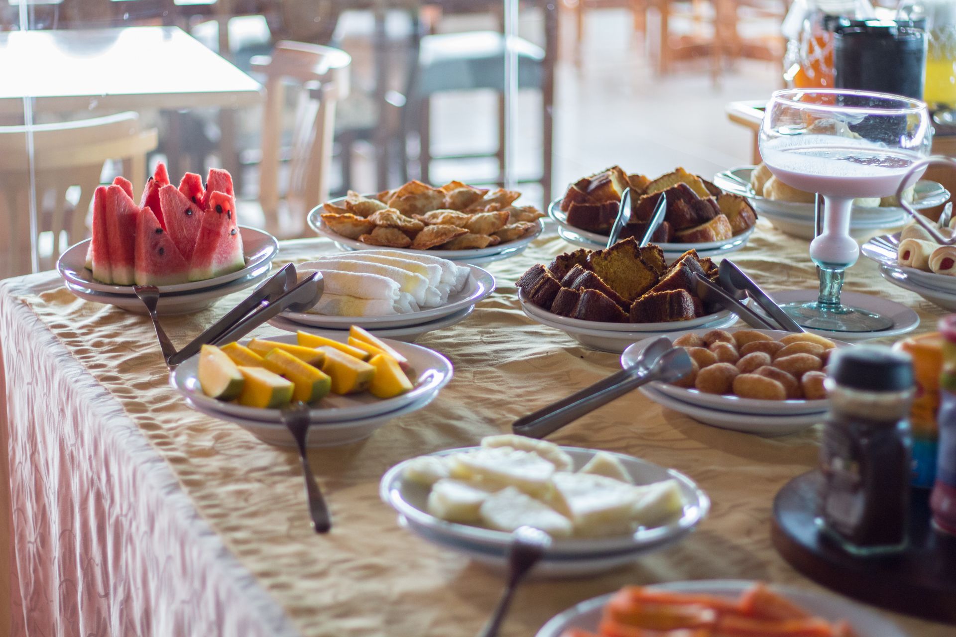 Mesa de buffet com diversos pratos, incluindo melancia, frutas, doces e uma bebida.