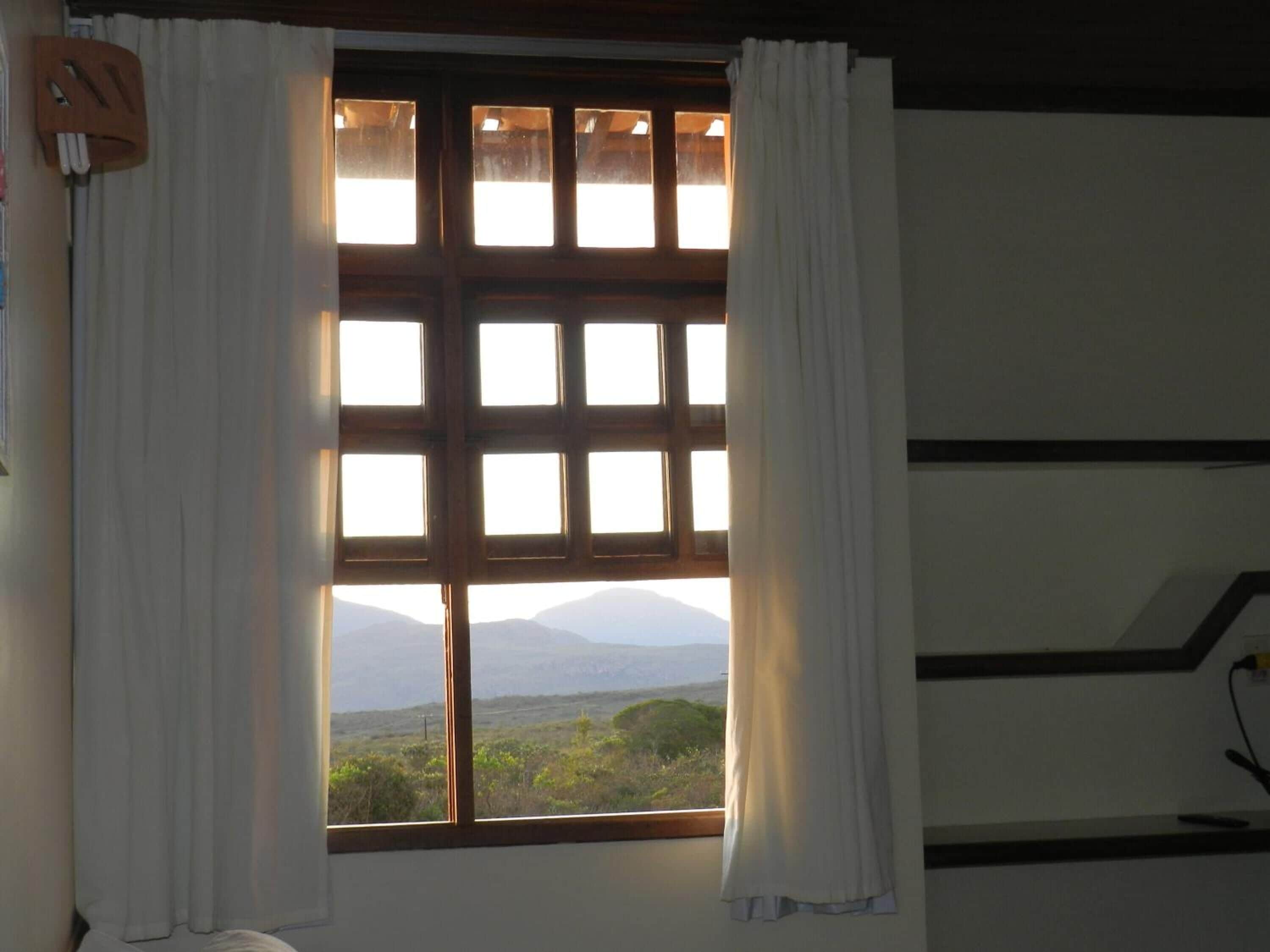 A room with a large window featuring a grid of panes, white curtains, and a view of a landscape with mountains.