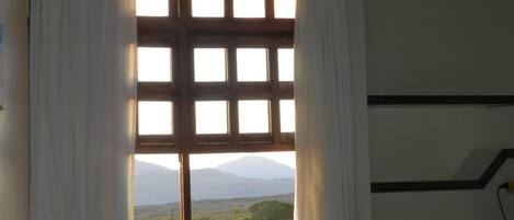A room with a large window featuring a grid of panes, white curtains, and a view of a landscape with mountains.