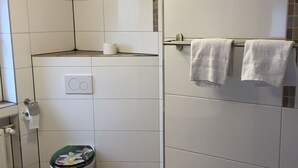 Un baño con azulejos blancos, un inodoro con cubierta floral, un perchero con dos toallas y suelo de madera.