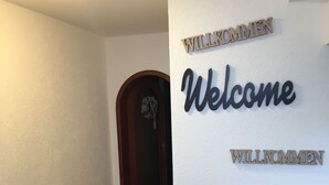 Un pasillo con piso de madera, una puerta adornada con un arreglo floral y una pared con letreros que dicen 'Welcome' en inglés y 'Willkommen' en alemán.