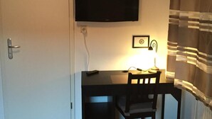 Una habitación con televisión en la pared, un escritorio con silla, una lámpara y un cuadro enmarcado.