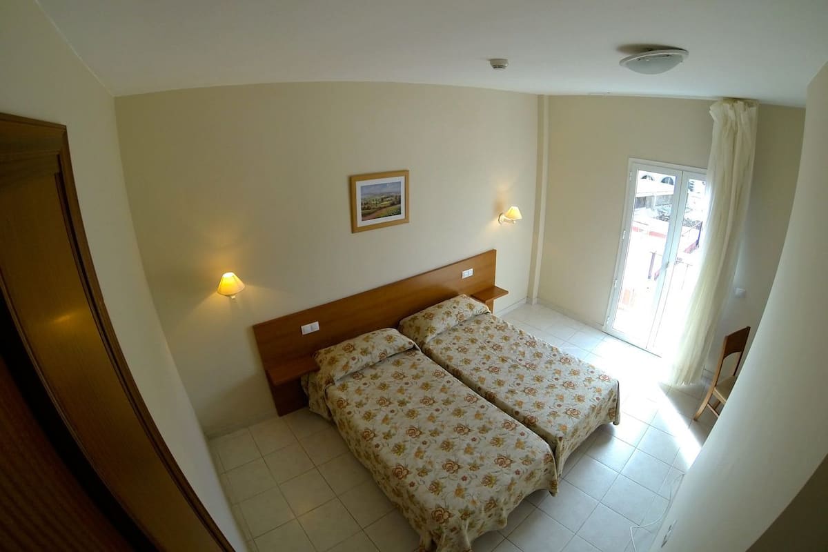 Une chambre avec deux lits, une tête de lit en bois, une petite fenêtre avec des rideaux et un tableau accroché au mur.
