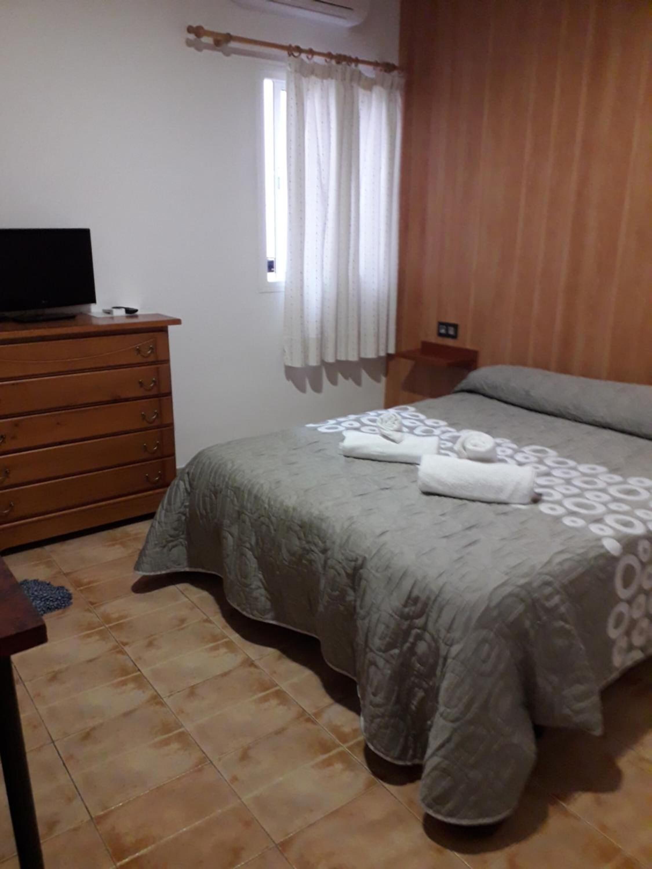 Une chambre à coucher avec un lit, une commode en bois, un téléviseur posé sur la commode et une fenêtre avec des rideaux.