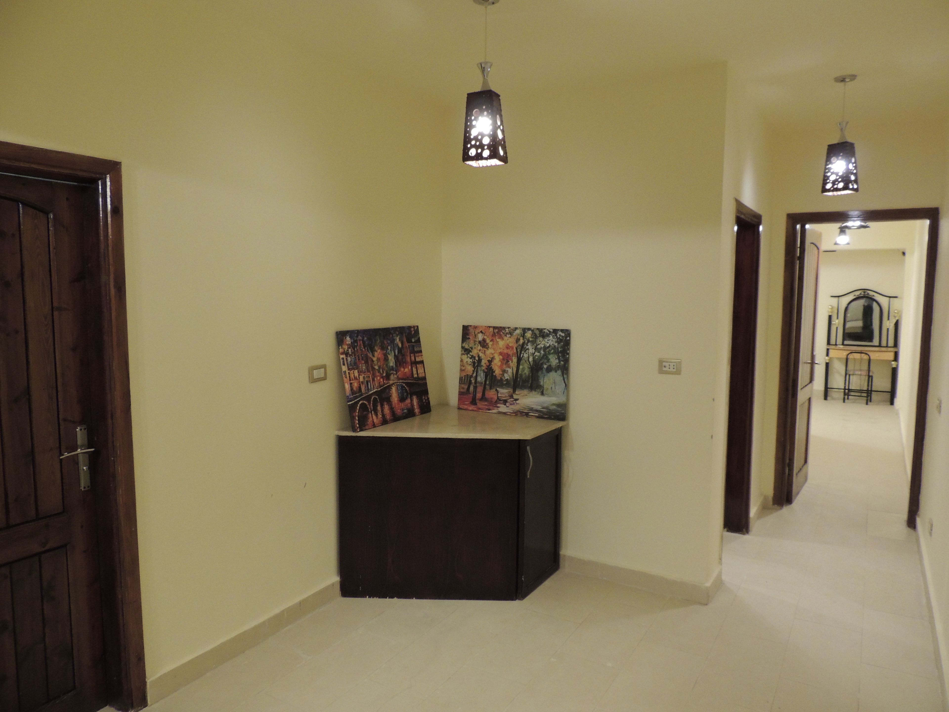Un couloir avec deux tableaux accrochés sur un meuble de couleur sombre, une porte en bois sur la gauche et, au fond, un bureau avec une chaise.