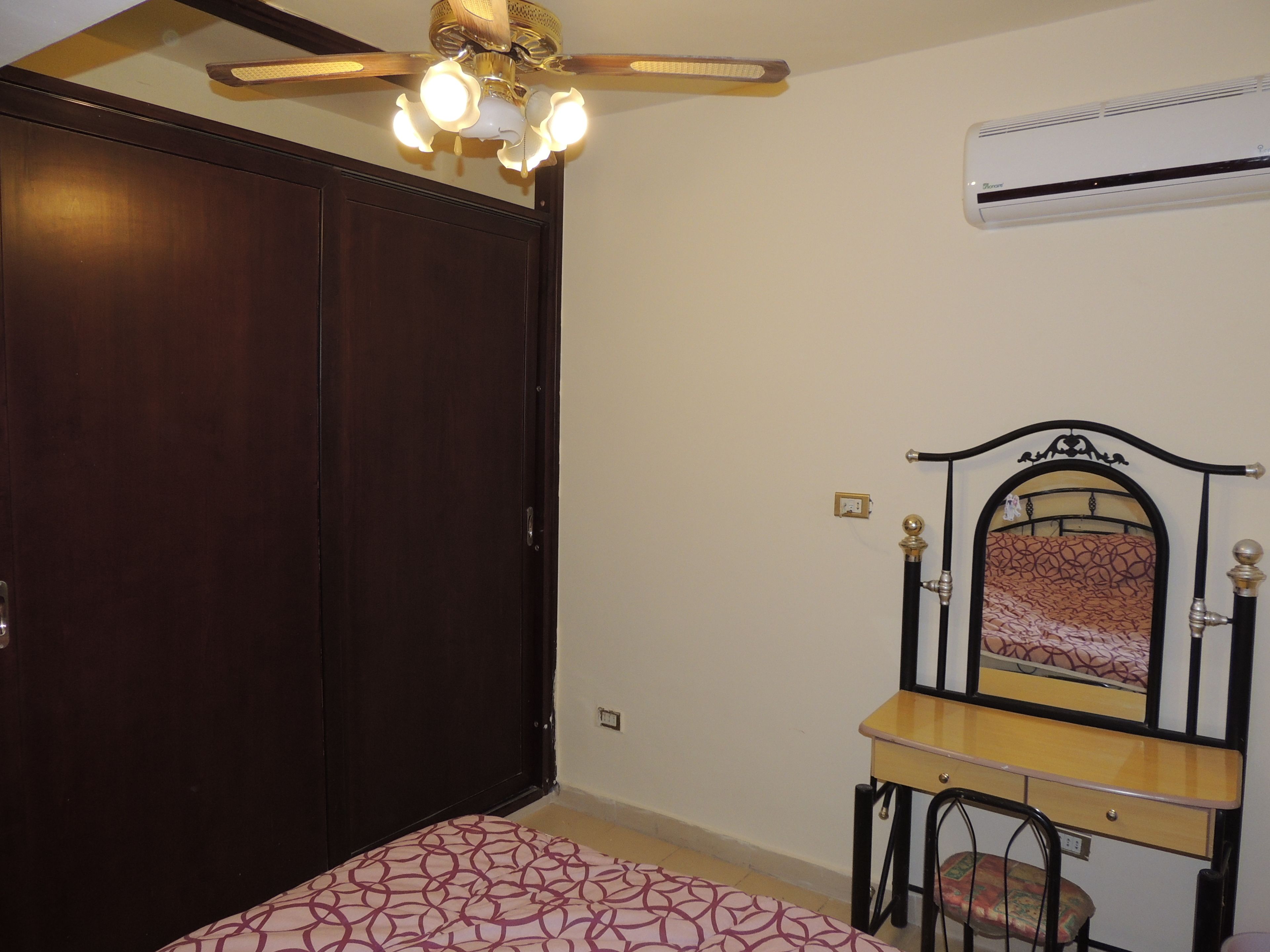 A bedroom with a bed, a wardrobe, a vanity with a mirror, and an air conditioning unit.