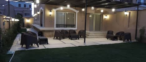 A covered outdoor seating area with multiple chairs and tables, a small staircase leading to a balcony, and wall-mounted lights.
