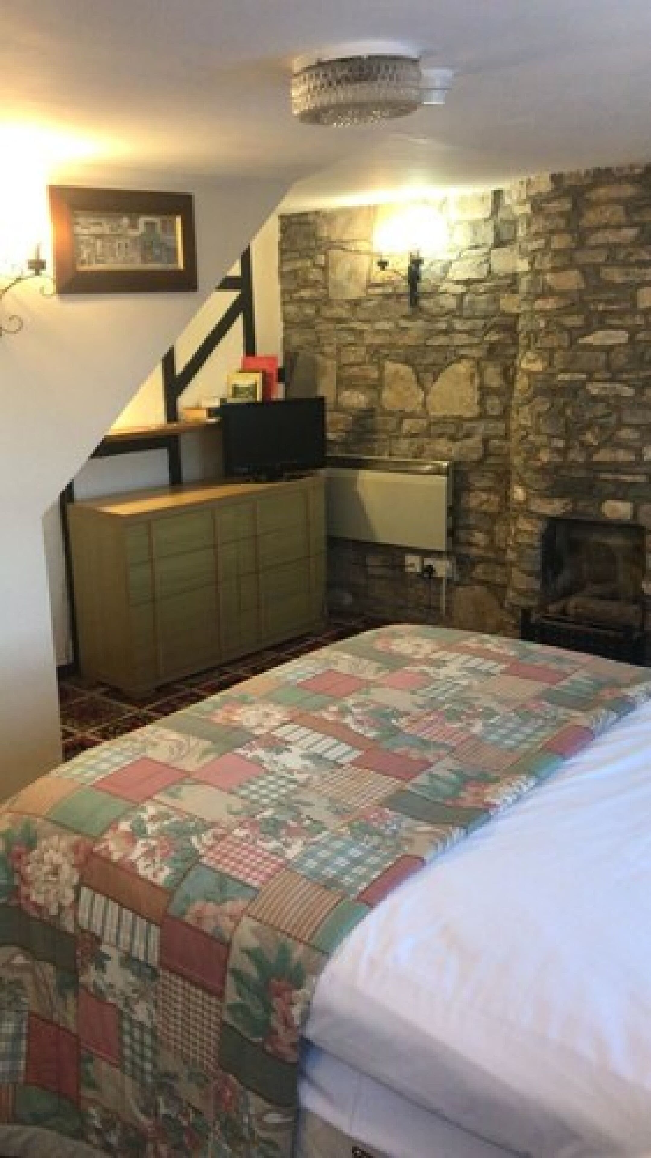 A bedroom with a stone wall, a wooden dresser, a quilted bedspread, and a staircase leading to an upper level.