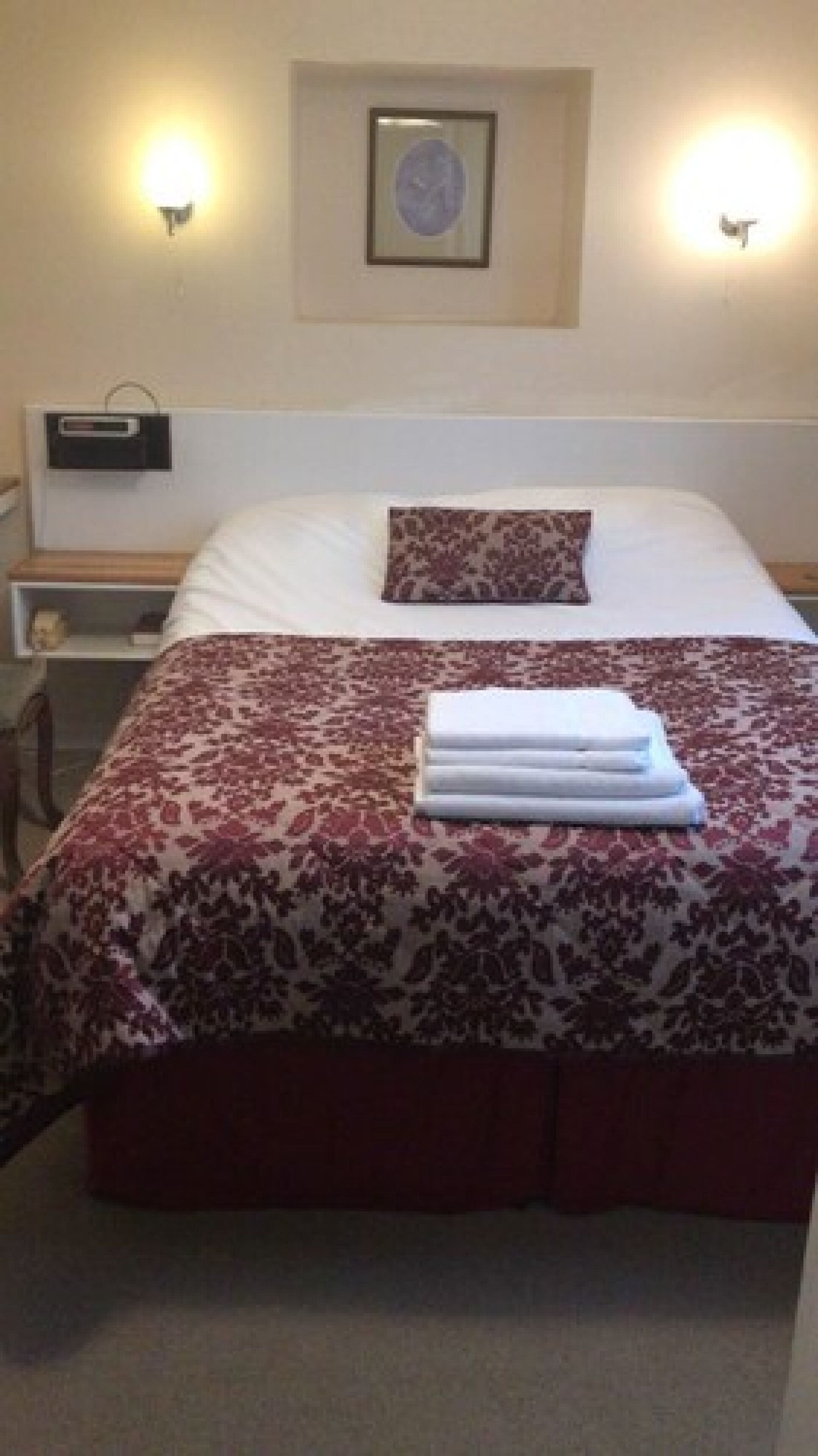 A neatly made bed with a patterned bedspread, a pillow, and a folded towel on the bed. There is a bedside table with a lamp and a small decorative item.