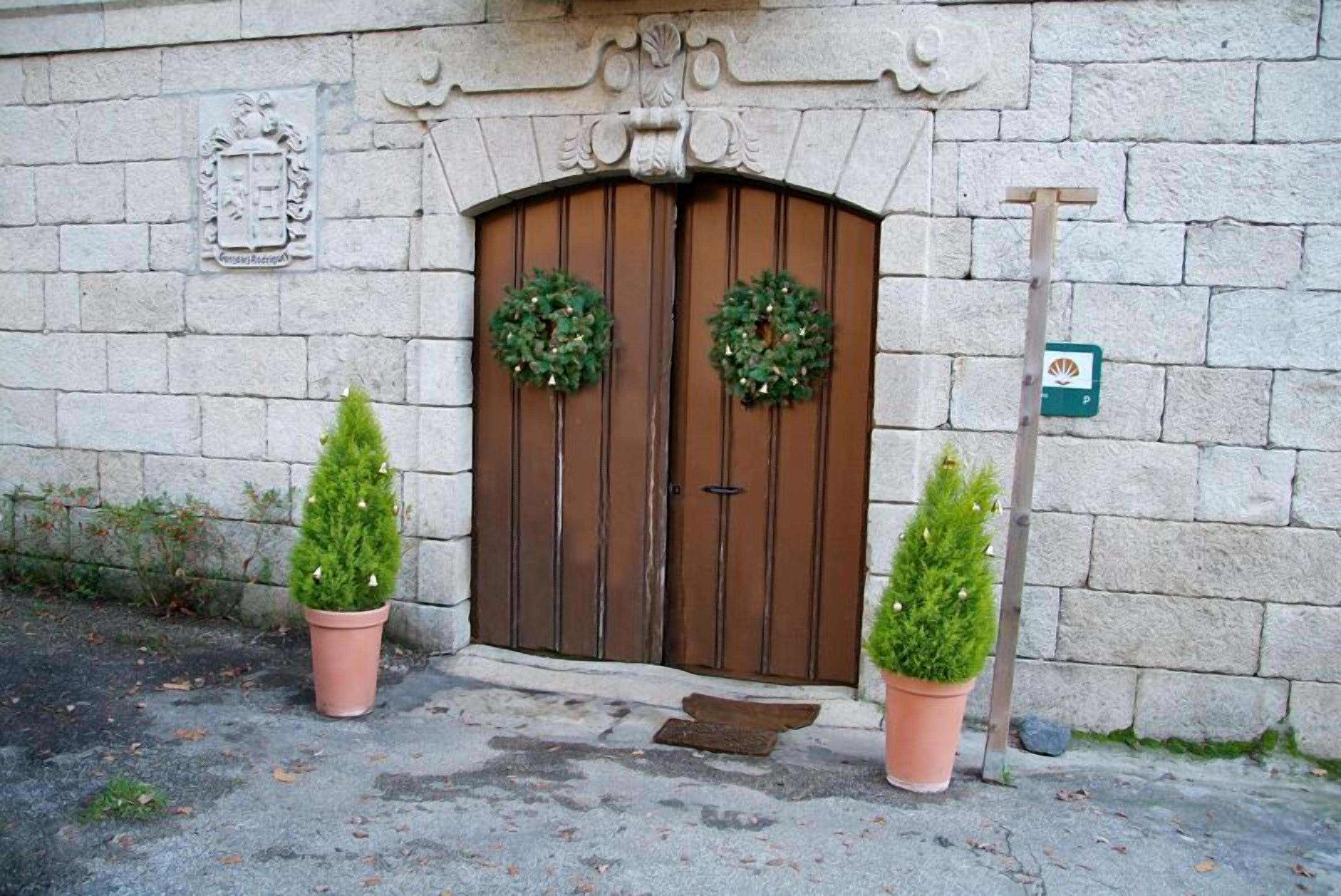 Un bâtiment en pierre avec une grande porte en bois, deux arbres à feuilles persistantes en pot et une petite boîte aux lettres.