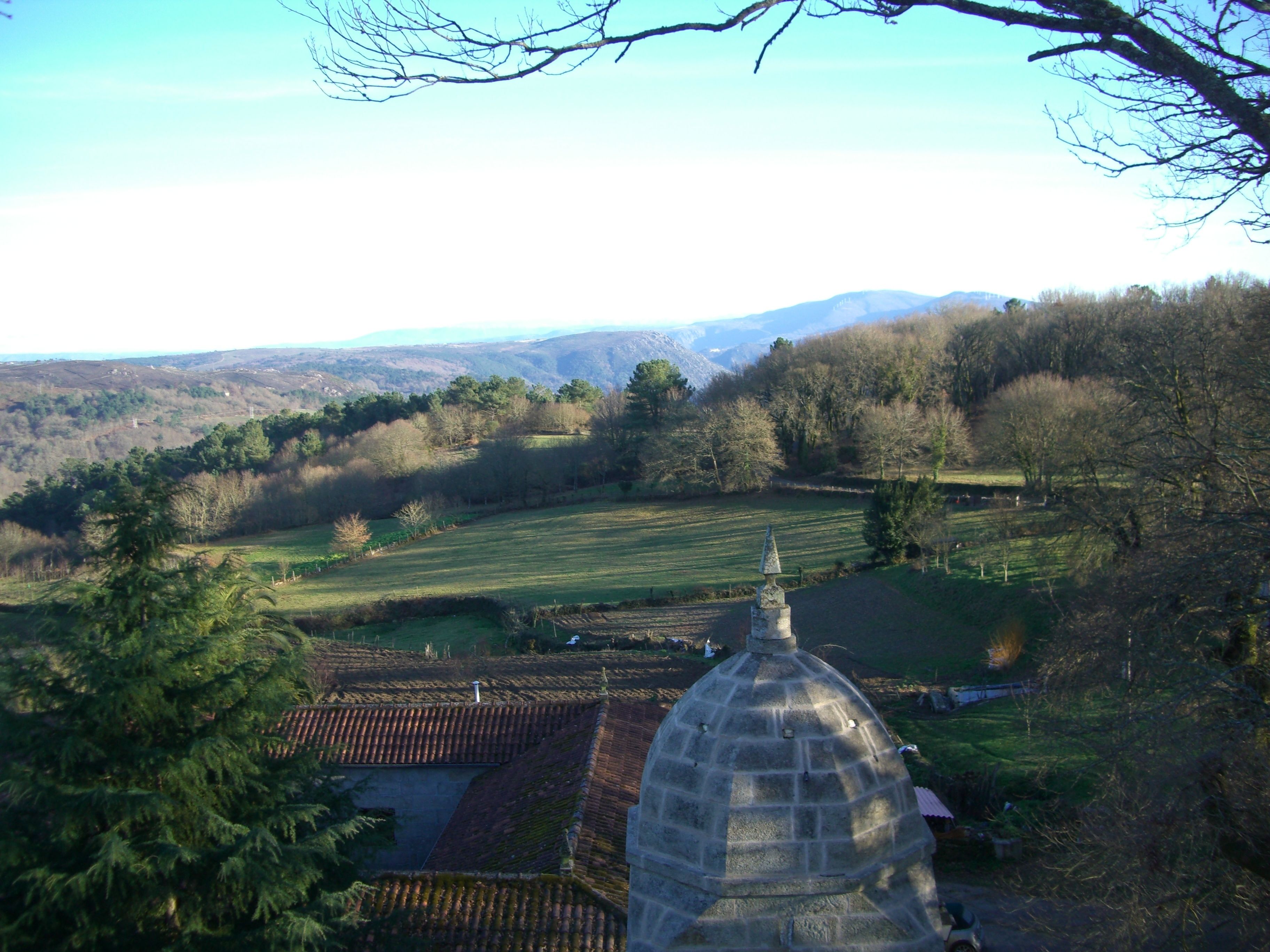 A rural landscape with a stone structure, possibly a chapel, surrounded by fields and trees.