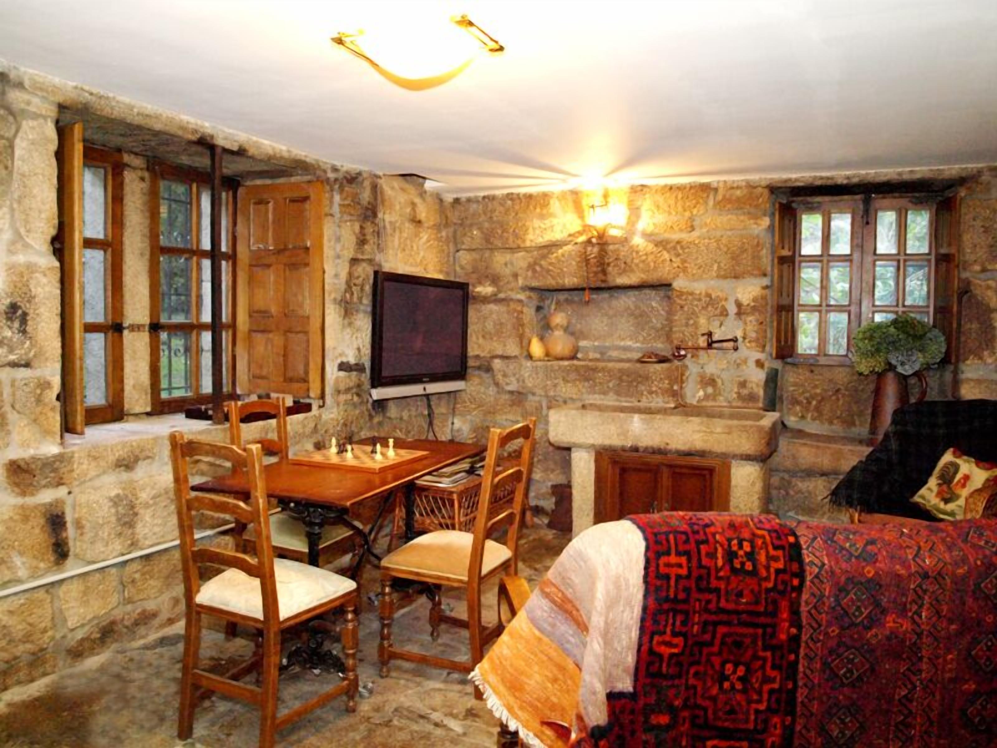 A stone-walled room with a fireplace, a television mounted on the wall, a chessboard on a wooden table, and a patterned throw on a sofa.