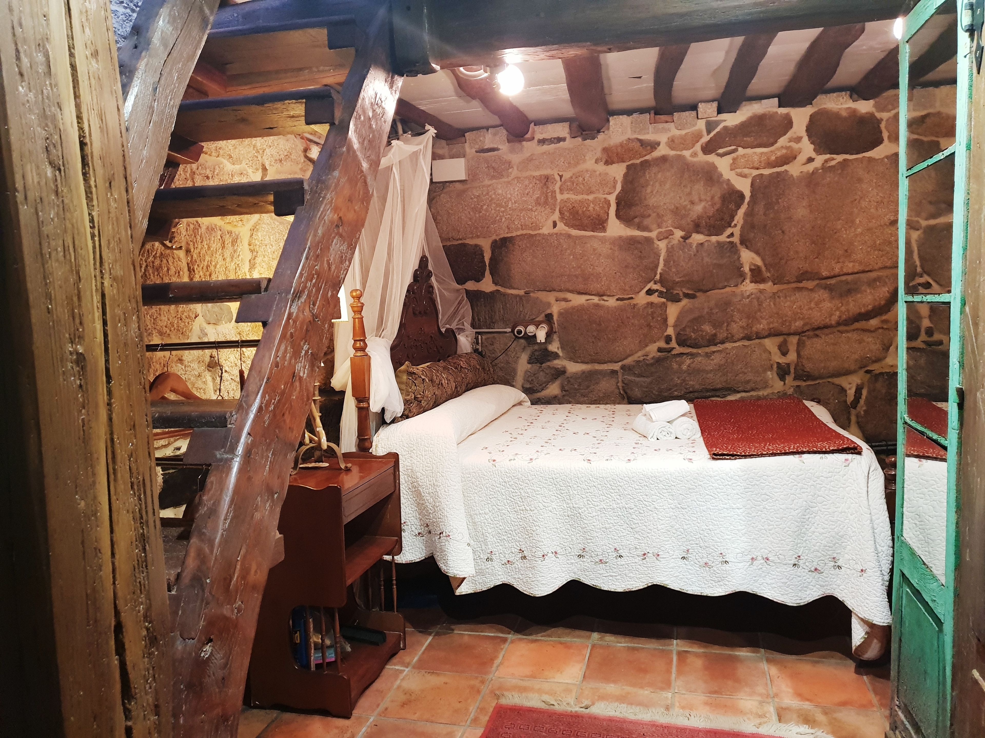 Une chambre à coucher au style rustique, avec un mur en pierre, des poutres apparentes, un lit recouvert d’une courtepointe blanche en dentelle et un plaid rouge.