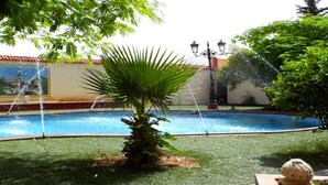 A swimming pool with a palm tree in the center, surrounded by a grassy area and a building with a tiled roof. - Hotel Bouazza (Tiaret)