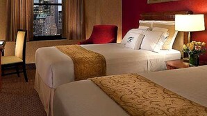 A hotel room with two beds, a desk, a chair, and a view of the cityscape. - Hotel Bouazza (Tiaret)