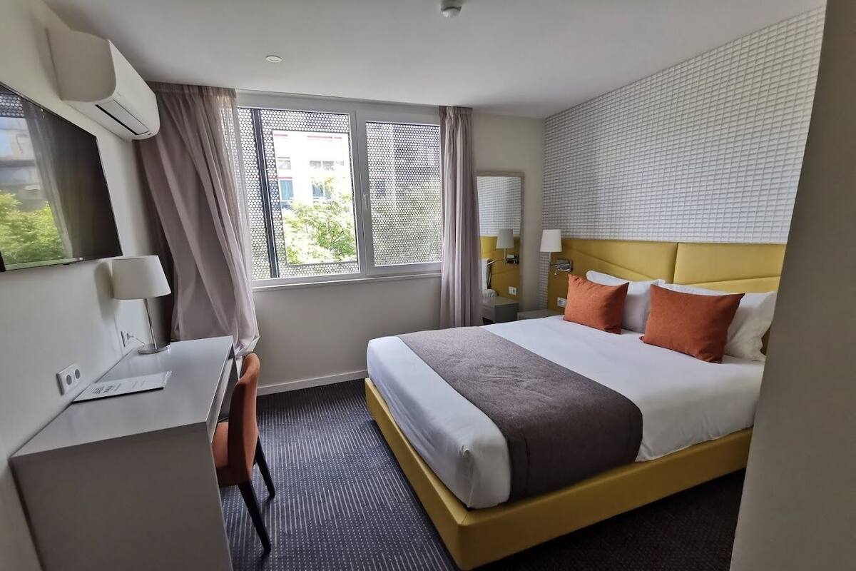 A hotel room with a bed, a desk, a chair, a window with curtains, and a wall-mounted air conditioner.