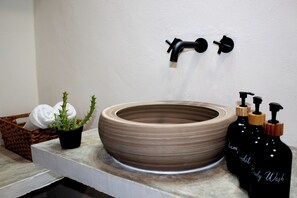 Casa de banho com uma grande pia redonda bege, duas torneiras pretas, uma pequena planta em vaso, uma cesta de vime com toalhas e dois frascos pretos rotulados "Chamondil Body Wash".