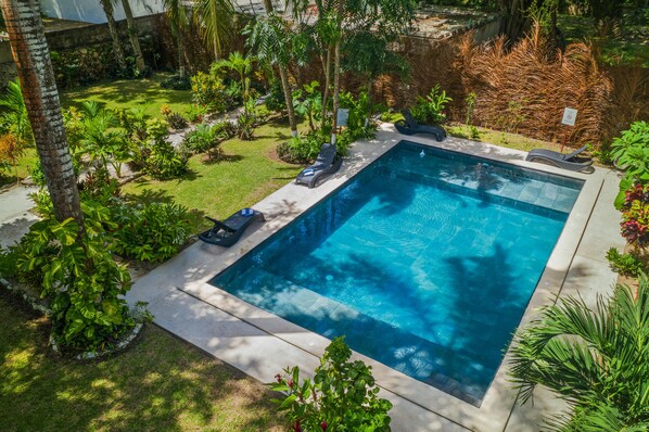 Uma piscina retangular com borda revestida, rodeada por um jardim bem cuidado com diversas plantas e árvores.
