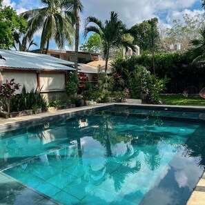 Piscina translúcida rodeada por vegetação exuberante e alguns edifícios.