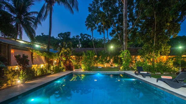 Ein Swimmingpool mit Liegestühlen, umgeben von tropischer Vegetation und einem strohgedeckten Gebäude.