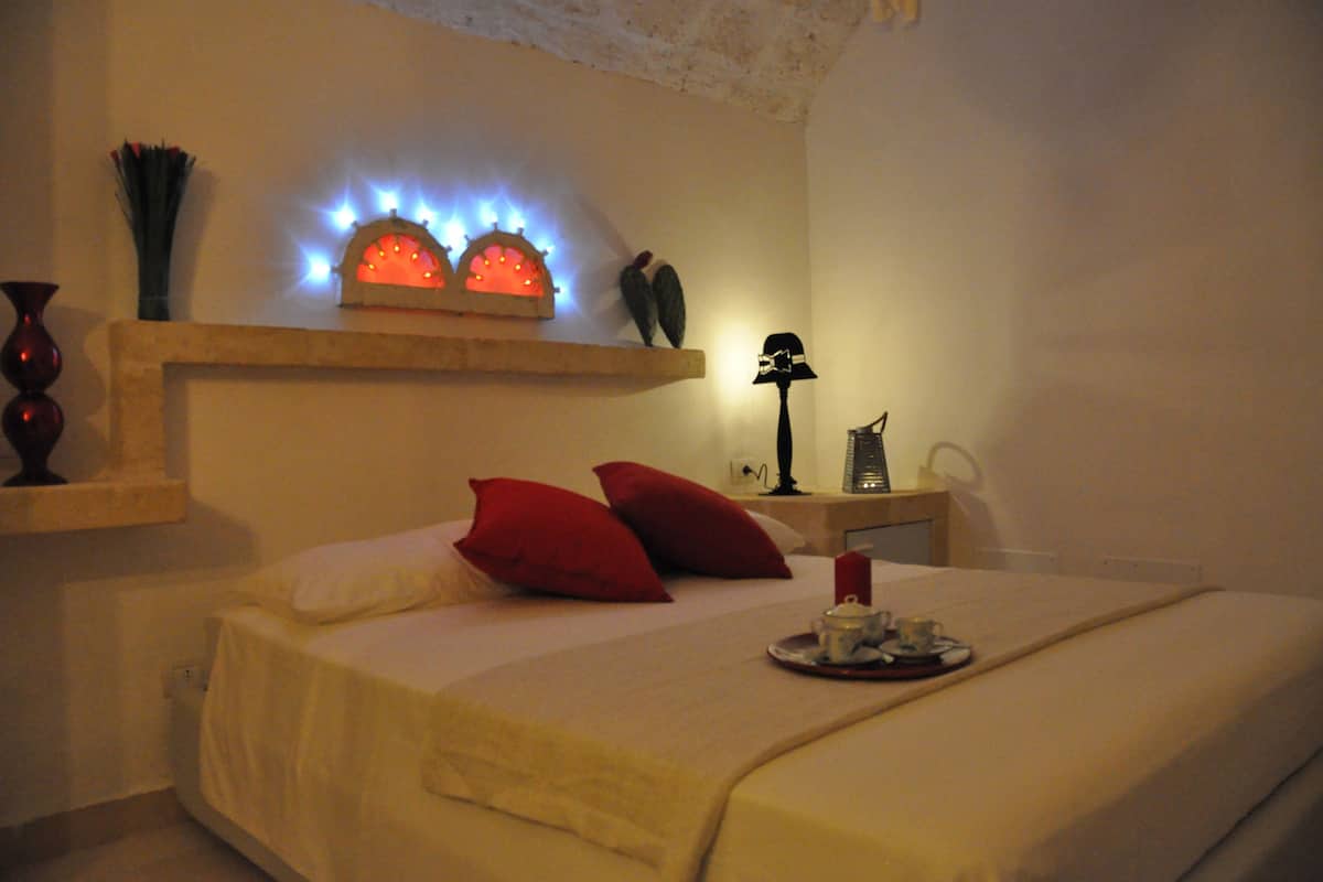 Hotellihuone, jossa on sänky, punaiset tyynyt, yöpöytä lampun kanssa ja seinälle kiinnitetty hylly, jossa on koriste-esineitä.