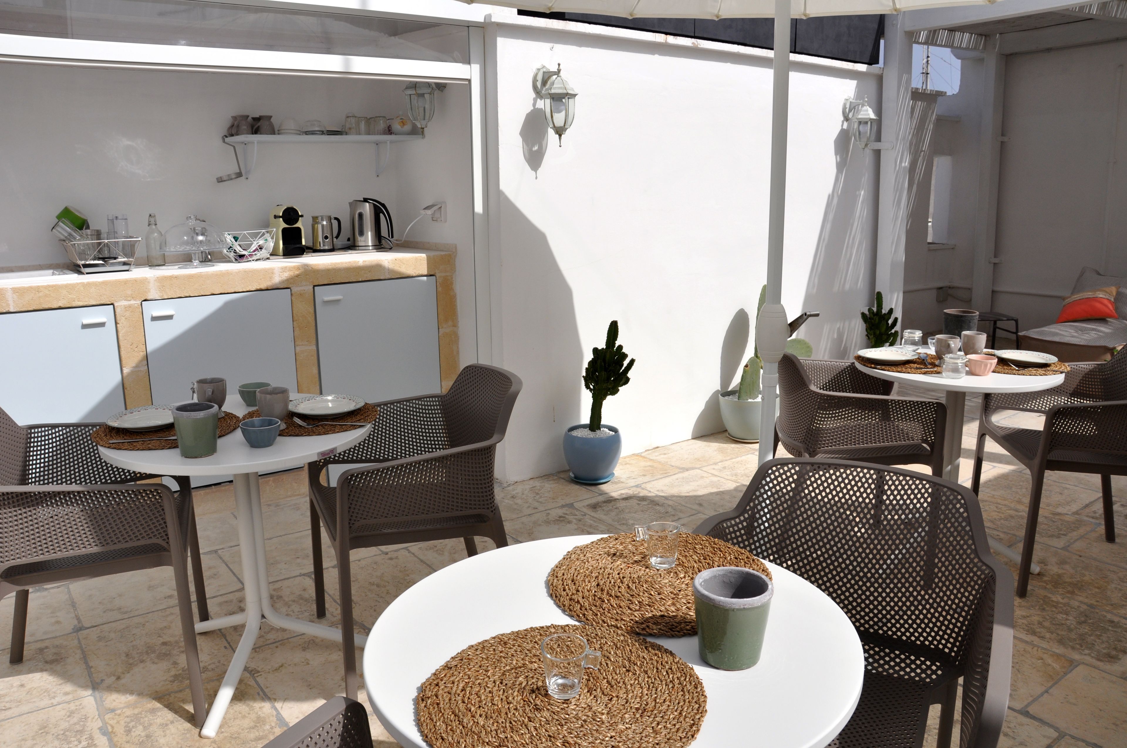 Un espace extérieur aménagé pour les repas, avec des chaises et des tables en osier, un coin cuisine doté d’un plan de travail blanc, et des plantes en pot.